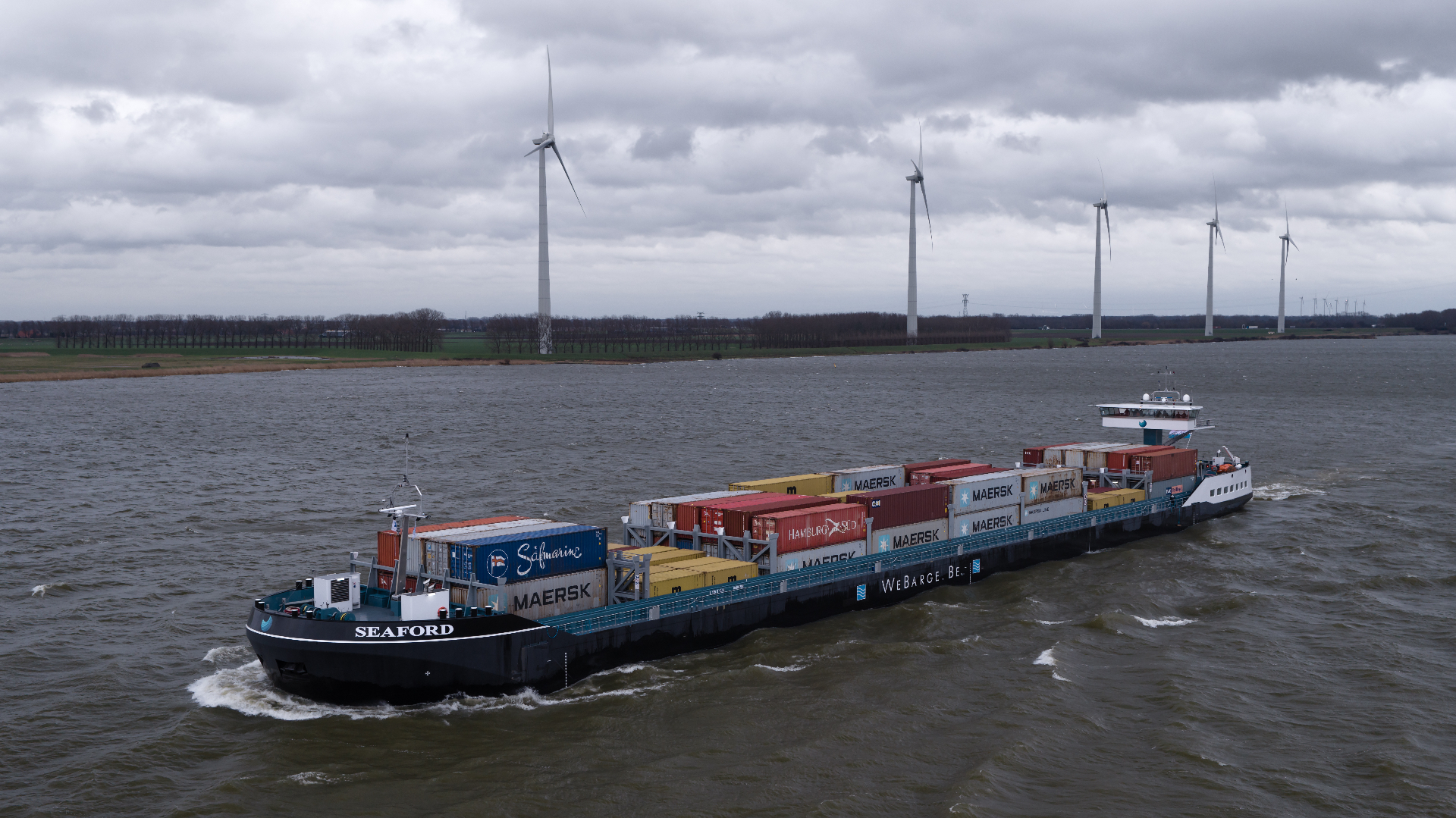 Inland container vessel Seaford underway past a wind farm